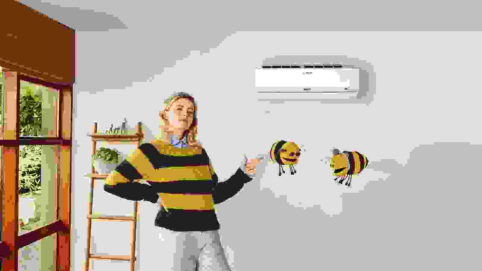 En kvinna i en gul- och svartrandig tröja pekar på en Bosch Climate 9100i luft-luftvärmepump som är monterad på väggen. Två tecknade bin svävar bredvid henne. Bilden är en del av kampanjen #LikeABosch